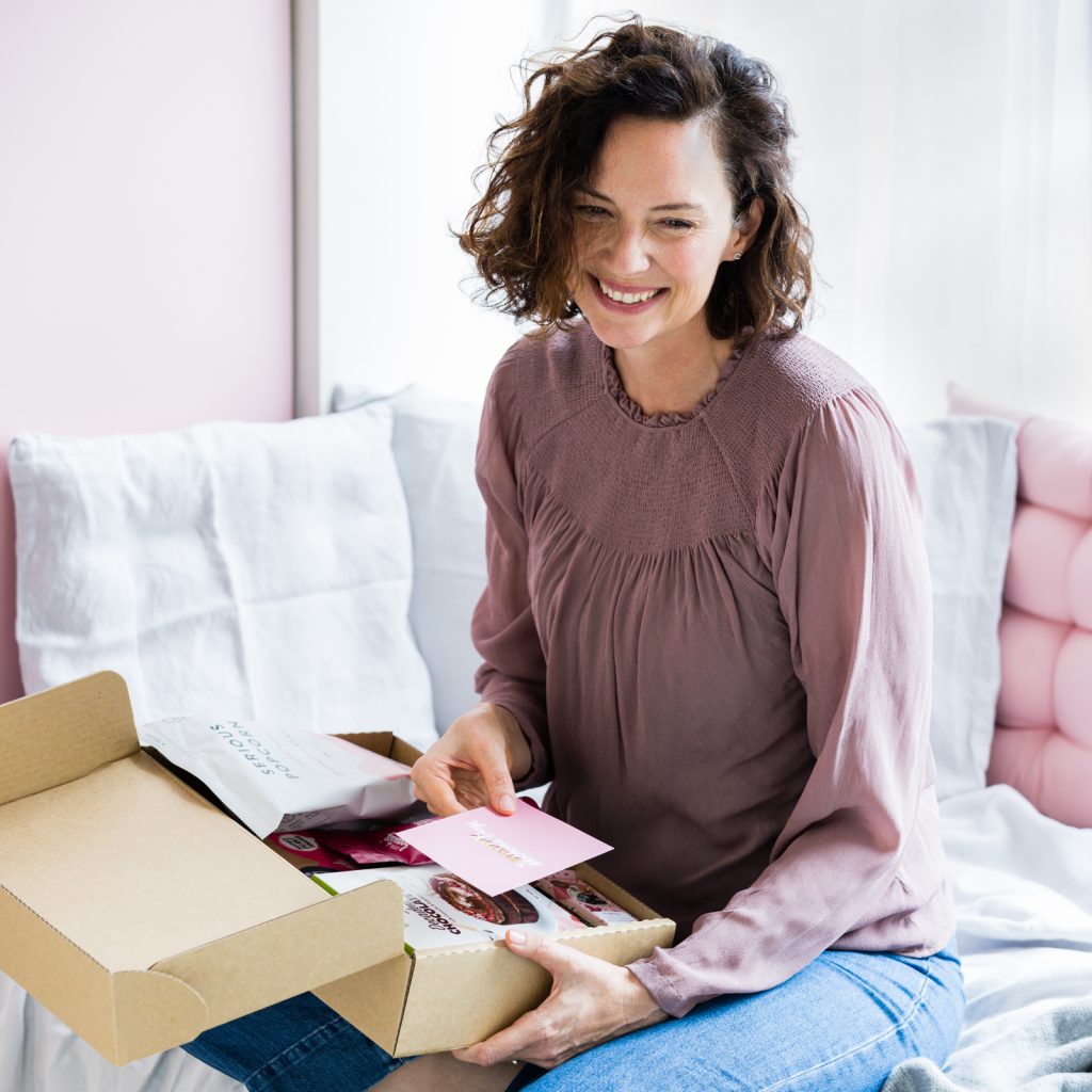 Women sits on couch smiling with a hamper of food on her lap.