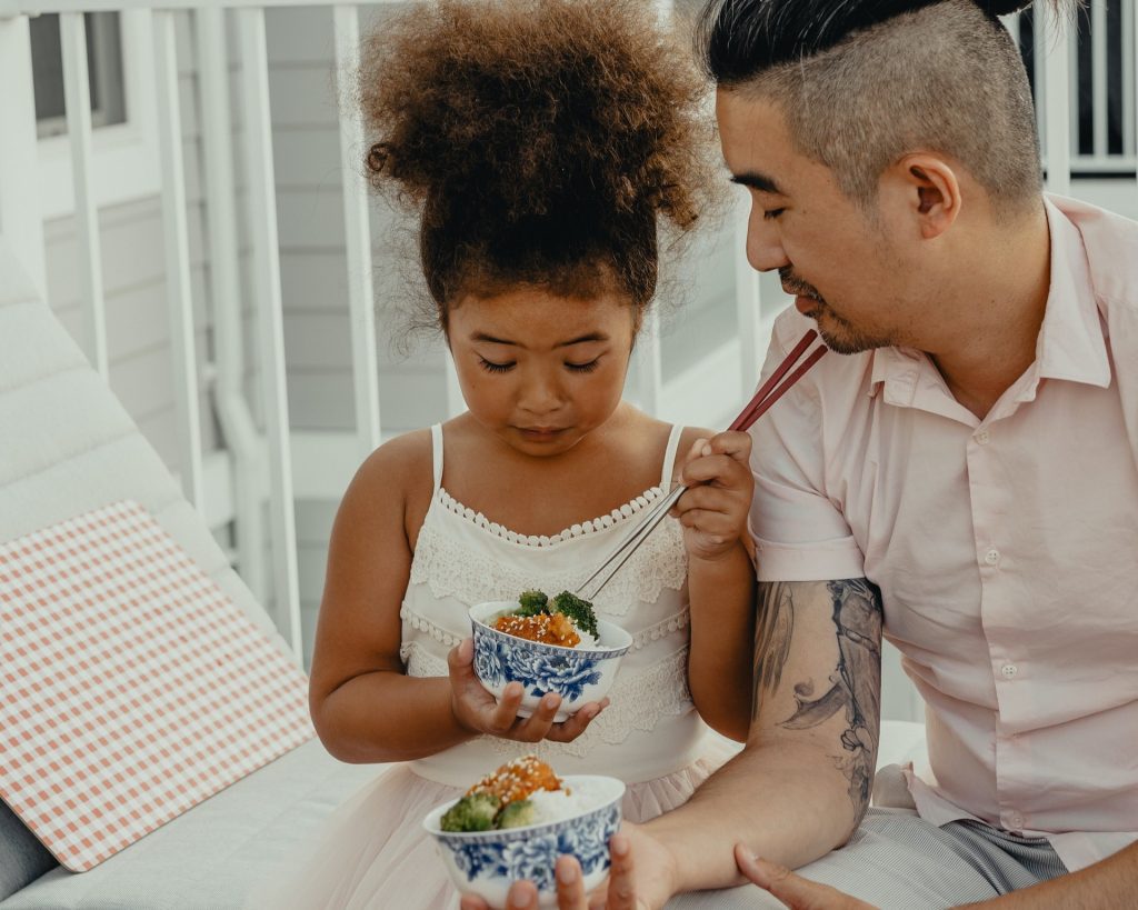 Dad and his daughter sit on white steps eating a bowl of rice together with chopsticks. Son looks at daughter while the daughter looks at her food