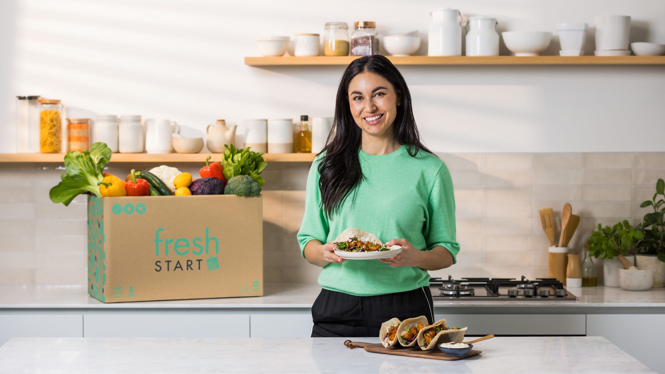 What Is the Healthiest Meal Kit in New Zealand? Your Guide to Nutrition-Focused Meal Deliveries 1 Nadia Lim holding healthy tacos standing next to a Fresh Start box packed with fresh vegetables and produce on a kitchen bench